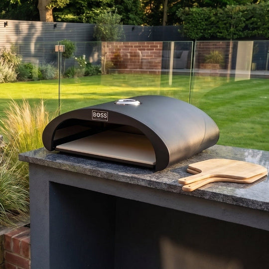 A dark gray outdoor pizza oven sits on a stone-topped built-in counter in a modern backyard setting with a green lawn, wooden deck, and garden plants.