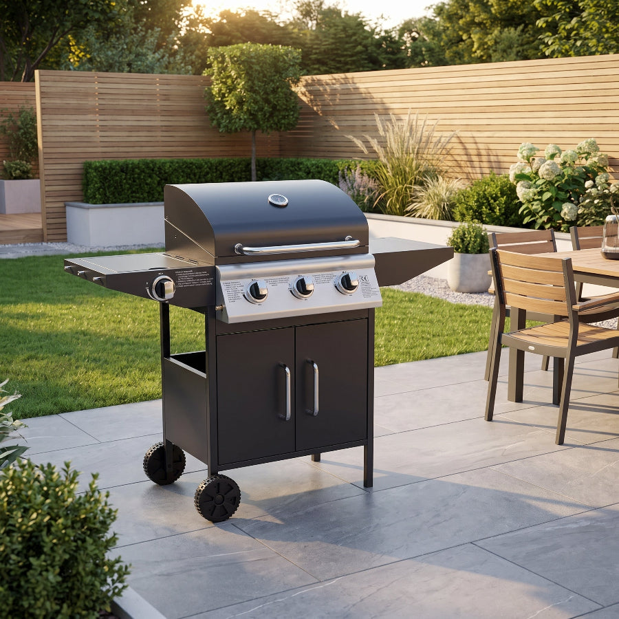 A black outdoor gas grill with a side table and lower storage cabinet, shown on a paved patio in a landscaped backyard with grass, plants, and a wooden fence.