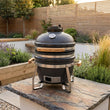 Load image into Gallery viewer, Outdoor setting with a large barbecue grill on a stone surface, surrounded by plants and wooden structures.
