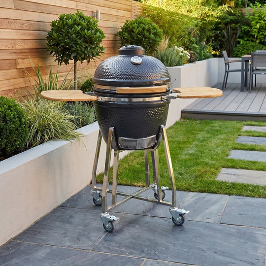 Black barbecue grill on wheels in a garden setting with greenery and a wooden wall.