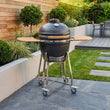 Load image into Gallery viewer, Black barbecue grill on wheels in a garden setting with greenery and a wooden wall.
