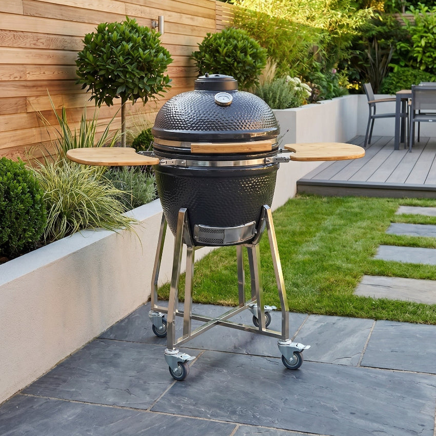 Black barbecue grill on wheels in a garden setting with greenery and a wooden wall.
