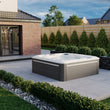 Load image into Gallery viewer, Outdoor hot tub in a garden setting with a brick building and trees in the background.
