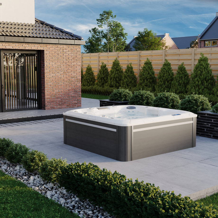 Outdoor hot tub in a garden setting with a brick building and trees in the background.
