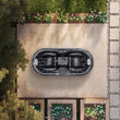 Load image into Gallery viewer, A top-down view of a black multi-seat hot tub placed on a beige stone patio in a landscaped backyard with plants and trees.
