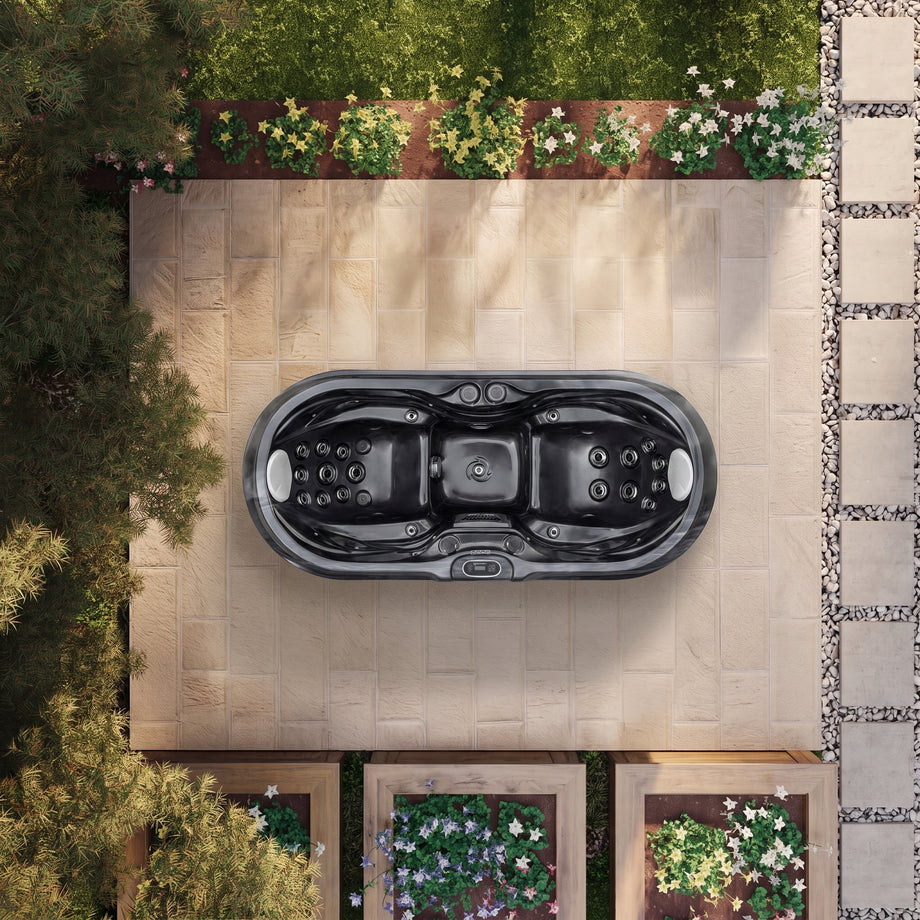 A top-down view of a black multi-seat hot tub placed on a beige stone patio in a landscaped backyard with plants and trees.