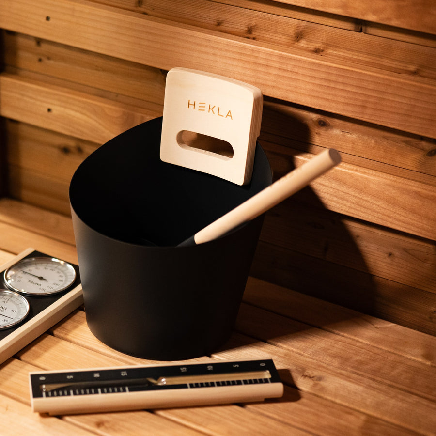 Hekla black aluminium sauna ladle resting in a black bucket on a timber sauna bench