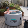 Load image into Gallery viewer, Woman relaxing in an inflatable ice pod outdoors on a wooden deck with plants and a building in the background.
